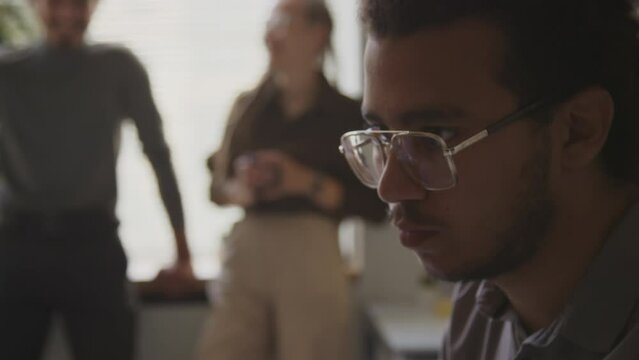 Tilting shot of focused Biracial male software engineer typing data in computer with keyboard while working in office with diverse colleagues chatting in blurred background
