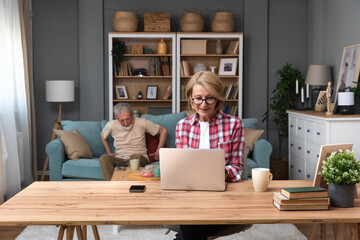 Senior woman sitting at desk with laptop computer watching online educational course. Older retired female video call with her friend while husband sitting in background on sofa. Modern simple living