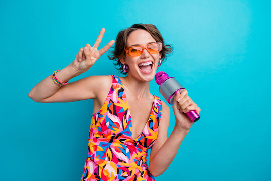 Photo of satisfied funky woman with bob hairdo dressed colorful clothes hold microphone show v-sign isolated on blue color background
