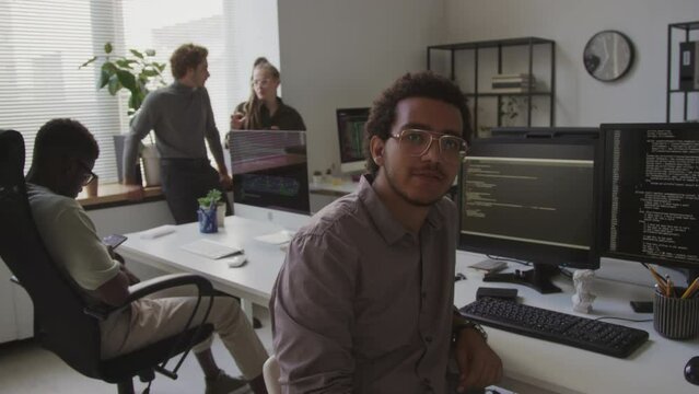 Medium portrait of young Biracial male programmer looking at camera while sitting in chair at workplace in modern open space office, working there with diverse colleagues