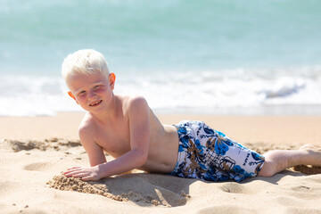 A smiling little boy lies on a beach against the backdrop of the sea and blue sky. Little boy lying on the sand and playing on the beach of Mediterranean sea. Summertime, vacation, travel banner.