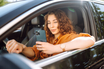 Young woman in casual clothes with a phone in her hands while driving a car. Happy woman uses a mobile application to rent a car. Concept of technology, transport.