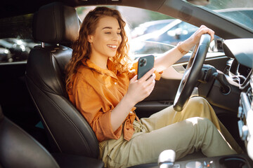 Young woman in casual clothes with a phone in her hands while driving a car. Happy woman uses a...
