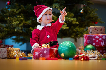 exciting one year old blond boy in red santa claus costume sitting with christmas hat and presents...