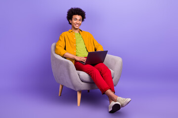Fototapeta na wymiar Photo of positive cheerful man wear trendy clothes sit chair use modern device netbook wifi connection isolated on purple color background