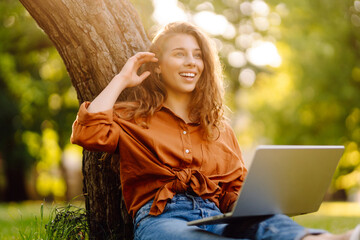 Beautiful woman in casual clothes on a green lawn with a laptop. Happy freelancer woman working on...