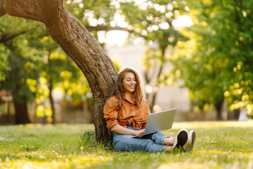 Beautiful woman in casual clothes on a green lawn with a laptop. Happy freelancer woman working on...