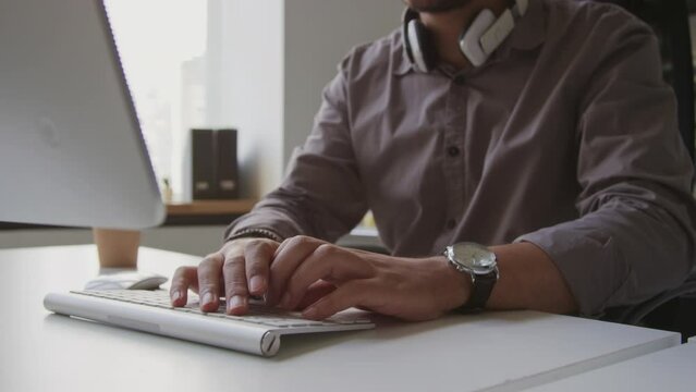 Cropped shot of unrecognizable male software developer typing on white wireless keyboard sitting in front of desktop computer at white office desk