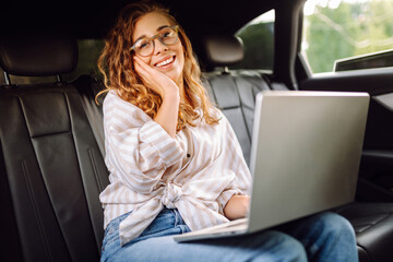 Beautiful business woman in casual clothes with a laptop working in the back seat of a car. Remote work concept, transport, technology.