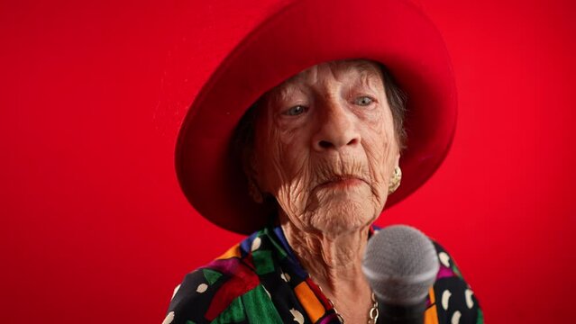 Funny fisheye view of elderly woman singing enthusiastically into a microphone and dancing wearing red hat isolated on red background