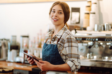 Portrait of a friendly female barista holding a tablet computer and smiling. A young coffee shop owner standing behind the bar using a digital tablet. Takeaway concept, technology.