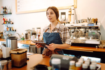 Portrait of a friendly female barista holding a tablet computer and smiling. A young coffee shop owner standing behind the bar using a digital tablet. Takeaway concept, technology.