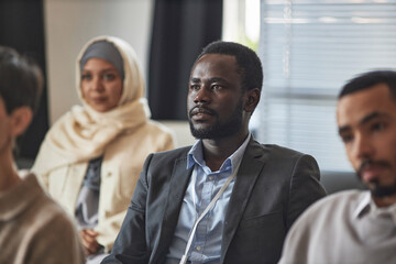Fototapeta na wymiar Young African American male employee in formalwear listening to speaker while sitting among multiethnic male colleague and Muslim woman