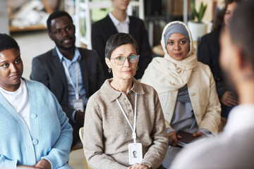 Group of serious intercultural employees looking at speaker making report or presentation and listening to him while sitting in lecture hall - obrazy, fototapety, plakaty