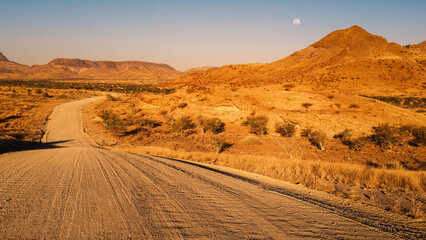 Warm sunset light bathing the Namib Desert traversed by a gravel road, east of Khorixas, Kunene,...