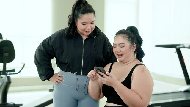 Overweight Asian woman sitting in gym, always exercise reduce belly, woman sits looks at food on phone female friend walks up invites come order food together, but friends complained.