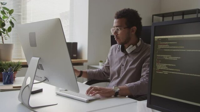 Waist up of concentrated Biracial male programmer typing on wireless keyboard while developing software on desktop computer, working in modern minimalist style office