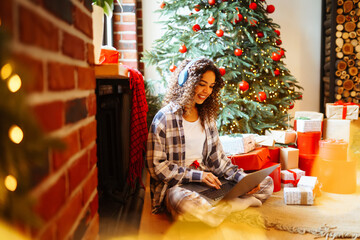 A beautiful woman uses a laptop at Christmas near the Christmas tree with gifts. Young woman makes online shopping near Christmas tree at home. Holidays concept, surprise.