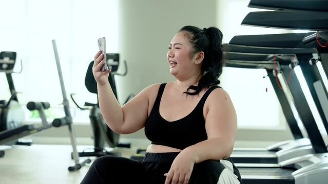 Chubby woman sits and rests after exercising in the fitness center. Video call with friends via smartphone To invite you to exercise. Happiness of a cute Asian overweight woman trying to lose weight