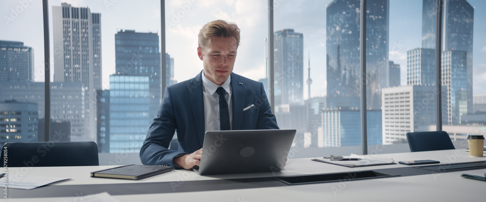 Poster confident businessman in a suit sitting at a desk in office, using laptop computer, next to window w