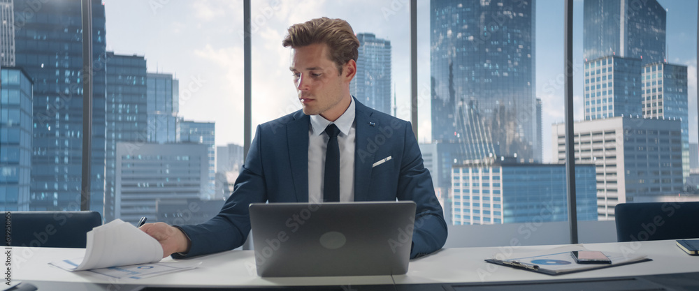 Poster confident businessman in a suit sitting at a desk in office, using laptop computer, next to window w