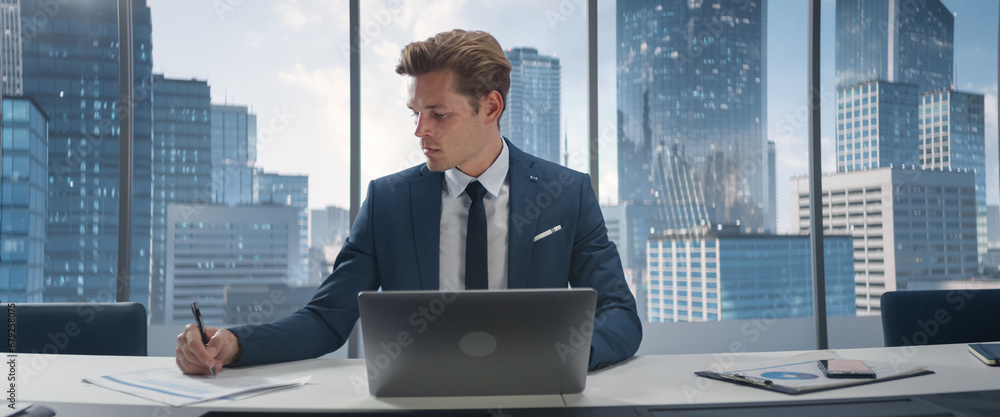 Poster confident businessman in a suit sitting at a desk in office, using laptop computer, next to window w