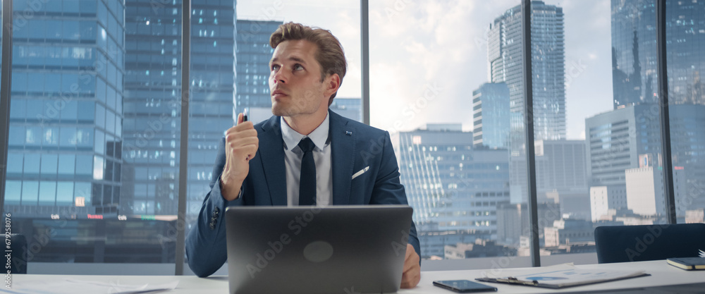 Poster confident businessman in a suit sitting at a desk in modern office, using laptop computer, next to w