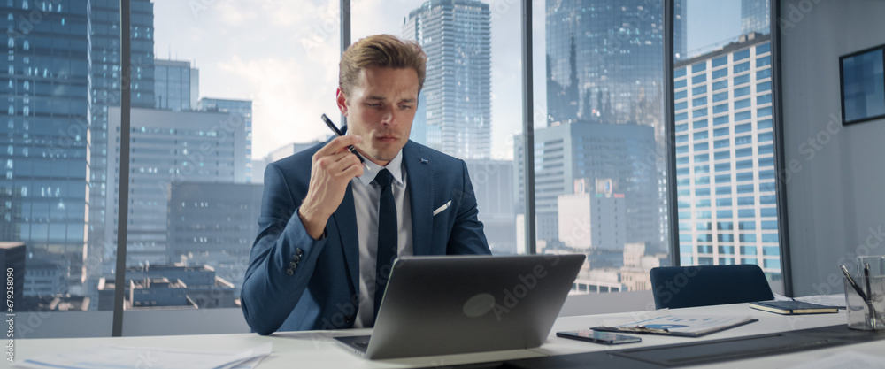 Poster confident businessman in a suit sitting at a desk in modern office, using laptop computer, next to w