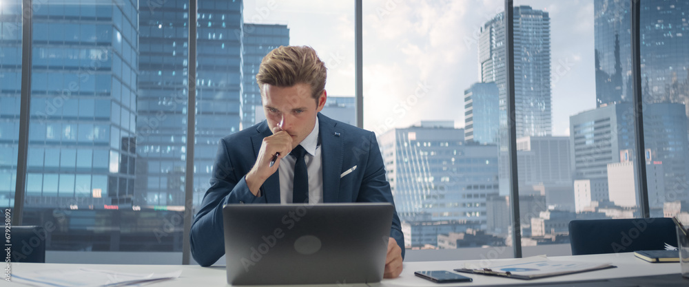 Poster confident businessman in a suit sitting at a desk in modern office, using laptop computer, next to w