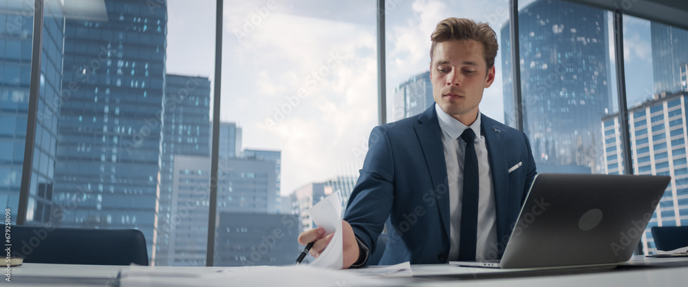Poster confident businessman in a suit sitting at a desk in modern office, using laptop computer, next to w