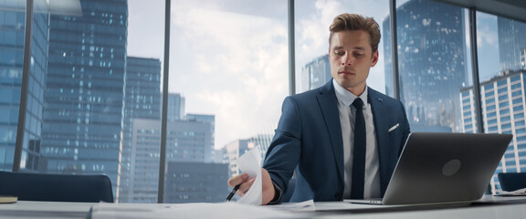 Confident Businessman in a Suit Sitting at a Desk in Modern Office, Using Laptop Computer, Next to...