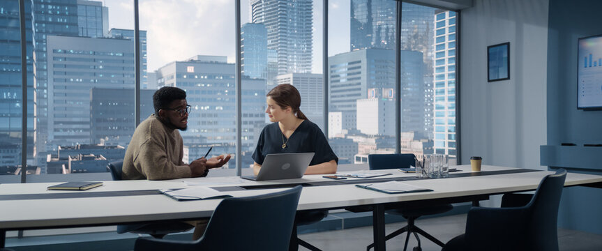 Two Multiethnic Colleagues Have a Discussion in Bright Modern Skyscraper Office in an Agency. Balck Creative Director and Female Project Manager Talking About Data On Laptop Computer.