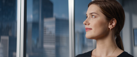 Close Up Beautiful Portrait of Caucasian Businesswoman Posing Next to Window in Big City Office with Skyscrapers. CEO Smiling, Looking At Camera. Successful Female Business Manager.