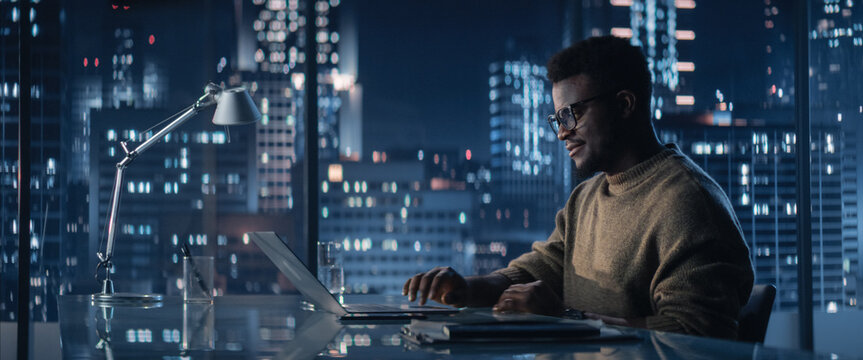Successful Handsome Black Businessman Working on Laptop Computer in Big City Office in the Evening. Financial Portfolio Manager Checking Data from Project Management Anual Report.