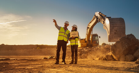 Caucasian Male Civil Engineer And Hispanic Female Architect Walking On Construction Site With Laptop Computer And Talking About New Real Estate Project. Machinery Working On Background. Golden Hour.