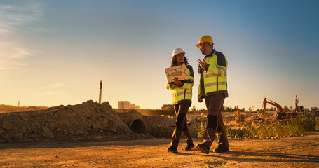 Caucasian Male Architect And Hispanic Female Urban Planner Walking On Construction Site With Laptop Computer And Talking About New Real Estate Project. Construction Equipment Working. Golden Hour - obrazy, fototapety, plakaty