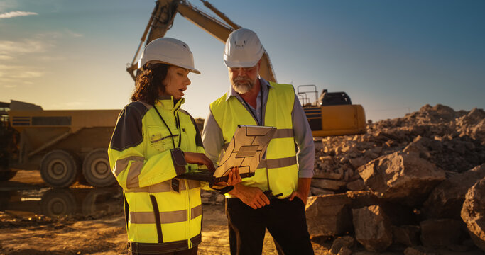 Caucasian Male Engineer And Hispanic Female Building Architect Walking On Construction Site With Laptop Computer And Talking About New Real Estate Project. Heavy Machinery Working. Golden Hour