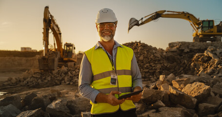 Successful Bearded Civil Engineer Wearing Protective Goggles And Smiling At Camera On Construction Site on a Sunny Day While Heavy Machinery Working. Man Holding Tablet, Wearing Hard Hat, Safety Vest. - obrazy, fototapety, plakaty