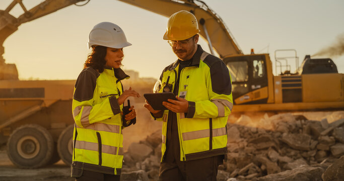 Cinematic Golden Hour Shot Of Construction Site: Caucasian Male Civil Engineer And Hispanic Female Inspector Talking, Using a Tablet. Trucks And Excavators Working On Land Development Project.