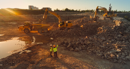 Aerial Drone Shot Of Construction Site On Sunny Evening: Industrial Excavators Digging Rocks And Loading Them Into A Truck. Engineer And Architect Observing Process, Discussing Real Estate Project - obrazy, fototapety, plakaty