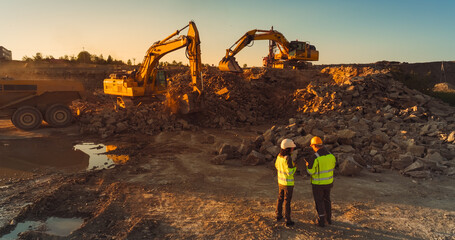 Aerial Drone Shot Of Construction Site On Sunny Evening: Industrial Excavators Digging Rocks And Loading Into A Truck. Civil Engineer And Inspector Observing Process, Discussing Real Estate Project - obrazy, fototapety, plakaty