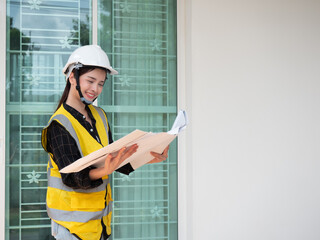 Portrait of woman engineer at building site smiling and looking at blueprint paper. Young construction manager standing in yellow safety vest and white hardhat.