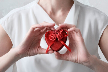 A woman in light clothes holds a red metal box in the shape of a heart with floral decoration at chest level. Conceptual gift, holiday