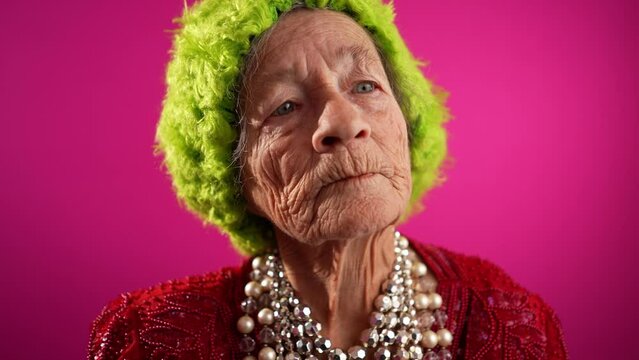 Slow motion funny view of mature elderly woman, 80s, having giving OKAY and thumbs up gesture, wearing green wig or hat isolated on pink background.