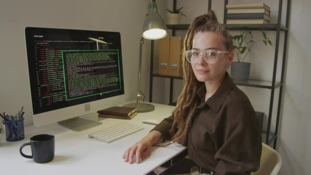 Waist up portrait of young attractive Caucasian female programmer with long dreadlocks posing for camera while sitting by computer with program codes on black screen at office desk in daytime