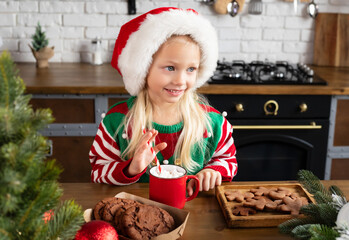 Kid smiling and looking sideways at wooden kitchen table with red mug of hot chocolate and marshmallow in. Child in Xmas sweater and Santa hat on Christmas decorated kitchen background