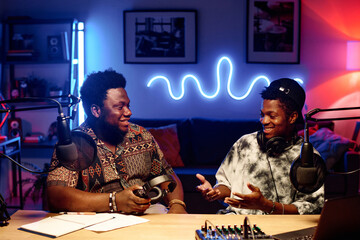 Two young African American men in stylish casualwear sitting in studio illuminated by neon lights...