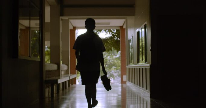 Silhouette scene while a male Asian high school student in white uniform with backpack walking on the corridor of his school and carrying his shoes for going back his home in the quiet evening.