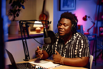 Young male radio presenter or host in stylish shirt speaking in microphone while sitting by desk in...