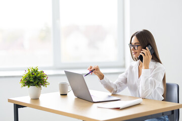 Cheerful young beautiful woman talking on the phone and looking at camera with smile while sitting at her working place